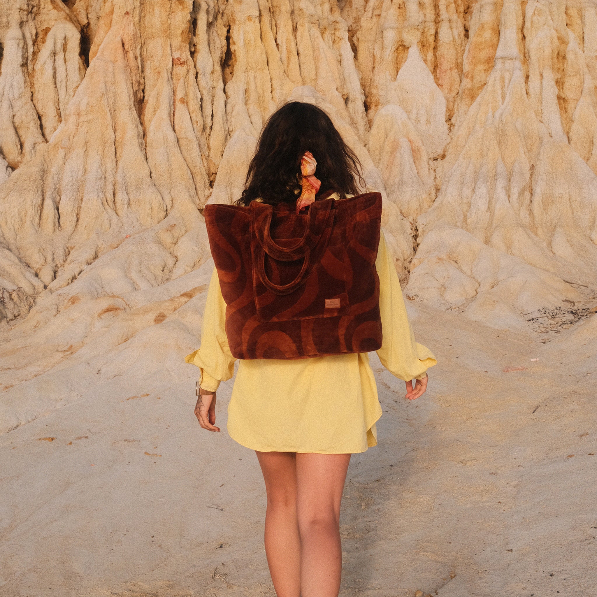 Person wearing a yellow dress and brown backpack walking in a desert-like landscape.