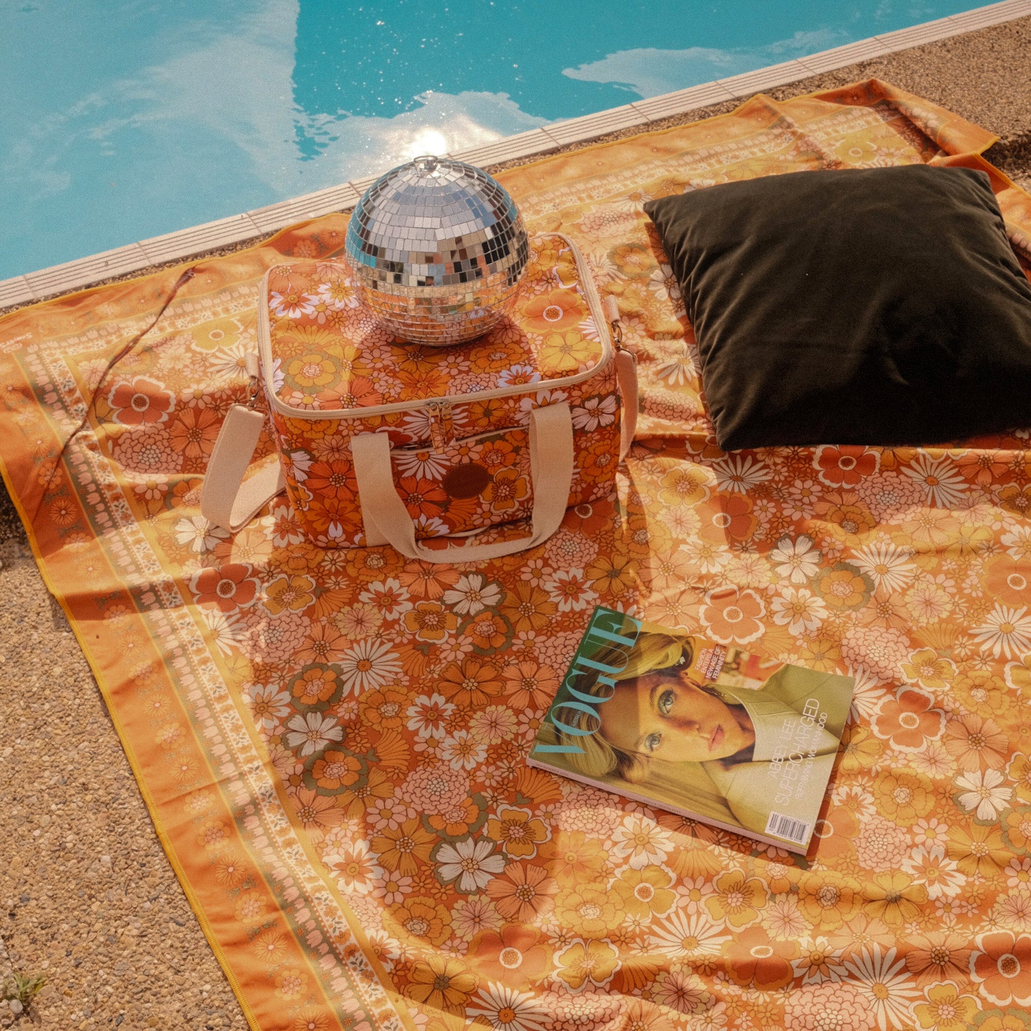 Floral-patterned bag with disco ball and magazine on a sunlit retro towel by a pool