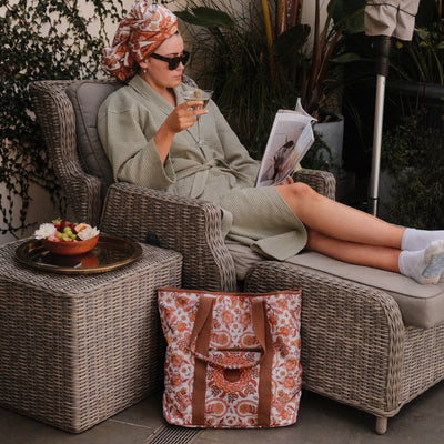 Woman in a robe reading a magazine on a wicker chair with a patterned bag beside her.