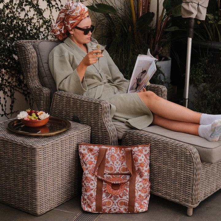 Woman in a robe reading a magazine on a wicker chair with a patterned bag beside her.