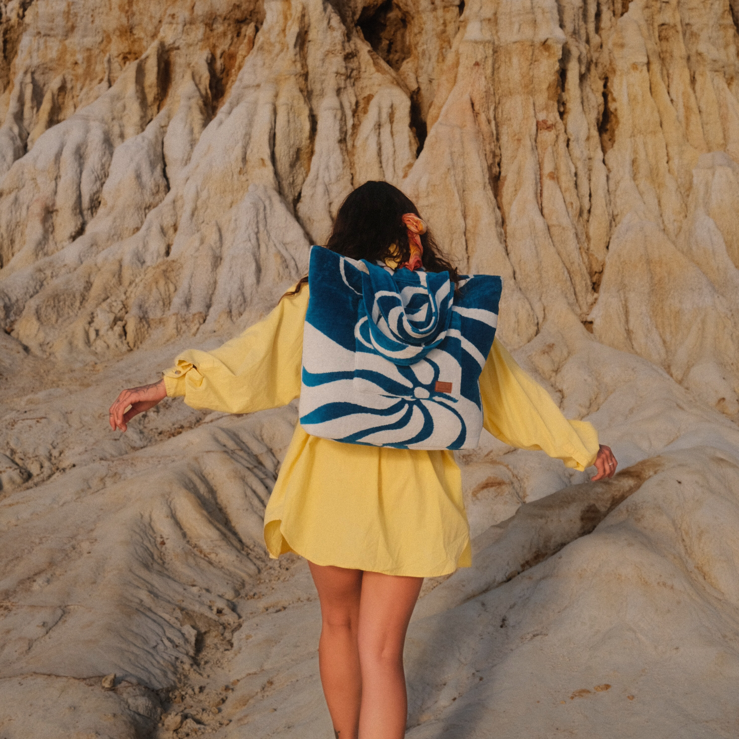 Person in yellow outfit with a blue and white patterned bag walking on sandy terrain.