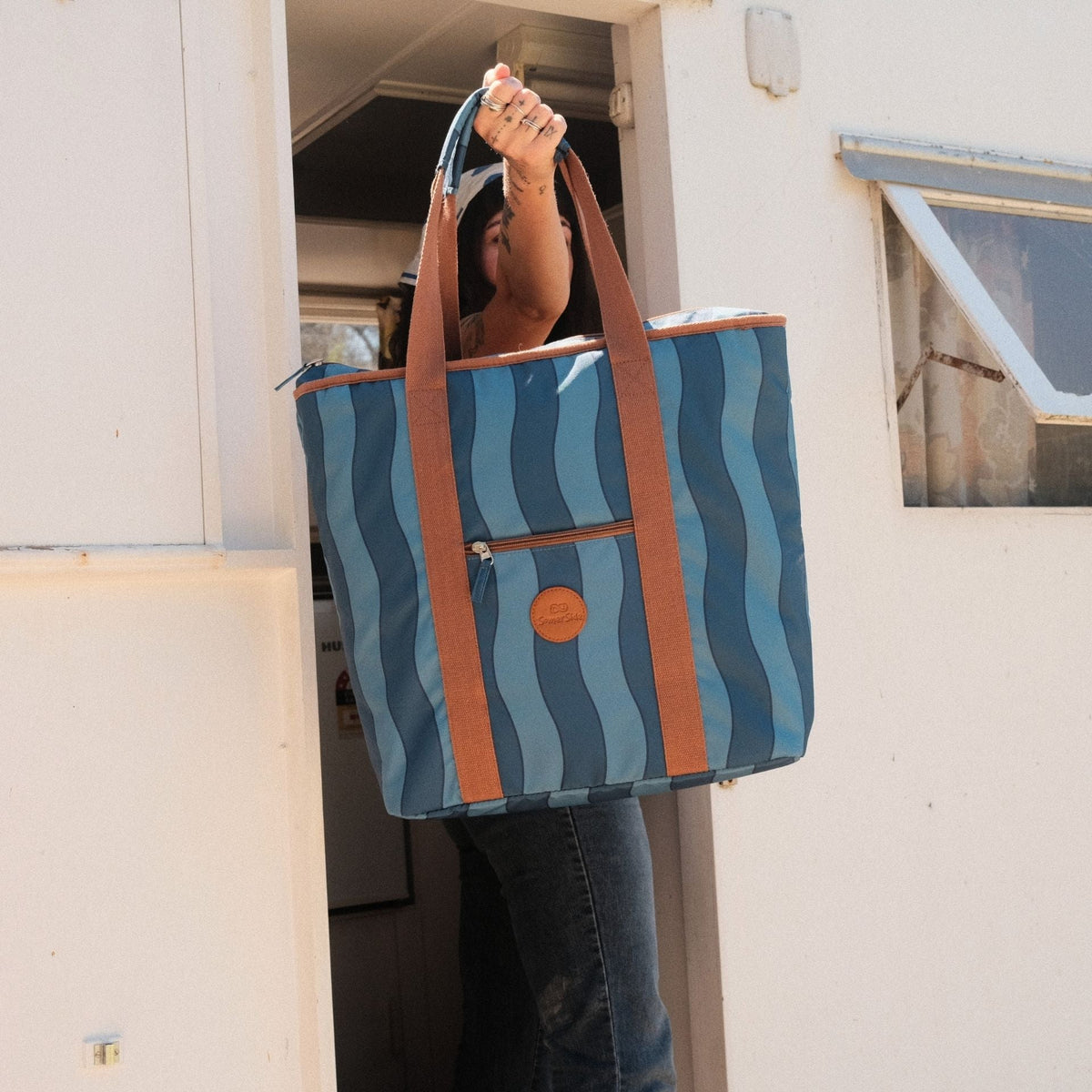 Jagger Tidal beach tote bag, two-tone blue wavy stripe, held up at a vintage caravan