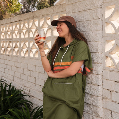 Stevie adult hooded towel poncho, olive green with retro orange stripes, leaning on a breeze block wall